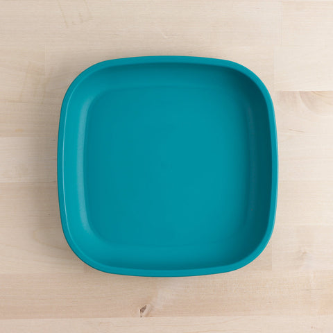 A Re-Play Flat Plate by REPLAY, featuring a square shape with slightly rounded corners in teal, crafted from recycled materials, is placed on a light wooden surface.
