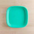 The Re-Play Flat Plate from REPLAY, designed as a square turquoise plate with rounded edges and made from recycled materials, is placed on a light wood surface. The plate's simple design and vivid color contrast beautifully against the natural, neutral background, exemplifying sustainable tableware at its finest.