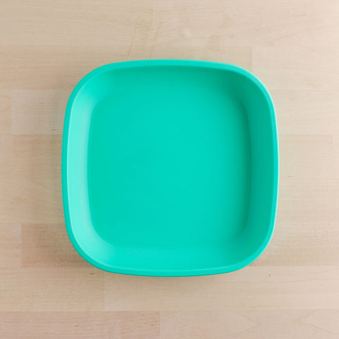 The Re-Play Flat Plate from REPLAY, designed as a square turquoise plate with rounded edges and made from recycled materials, is placed on a light wood surface. The plate's simple design and vivid color contrast beautifully against the natural, neutral background, exemplifying sustainable tableware at its finest.