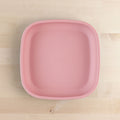 The REPLAY Re-Play Flat Plate is a square-shaped, pastel pink plastic plate featuring rounded edges. It rests on a light wood surface, showcasing its smooth, matte finish and medium size. Made from recycled materials, this sustainable tableware from REPLAY is both stylish and eco-friendly.
