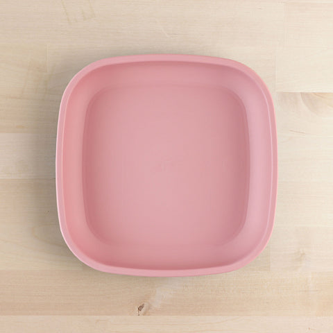 The REPLAY Re-Play Flat Plate is a square-shaped, pastel pink plastic plate featuring rounded edges. It rests on a light wood surface, showcasing its smooth, matte finish and medium size. Made from recycled materials, this sustainable tableware from REPLAY is both stylish and eco-friendly.