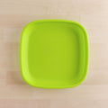 A bright green Re-Play Flat Plate from REPLAY, crafted in a square shape with rounded edges and made from recycled materials, rests on a light wooden surface. The durable plate is empty, and the image is taken from a top-down perspective.
