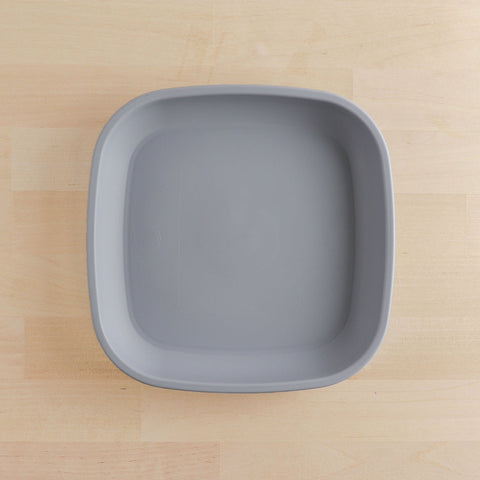 A top-down view of the REPLAY Re-Play Flat Plate, a square, light gray silicone plate with rounded corners placed on a light wooden surface. The plate, made from recycled materials, has a smooth and simple design, showing no additional features or textures.
