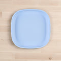 The REPLAY Re-Play Flat Plate, a square-shaped, light blue plastic plate made from recycled materials, is centered on a light wood surface. The plate features slightly rounded corners and smooth edges. The background showcases the natural grain pattern of wooden planks.