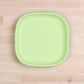 A Re-Play Flat Plate from REPLAY, crafted in a square shape with rounded corners and featuring a smooth finish, sits on a light wooden surface. This green plastic plate is made from recycled materials, embodying the principles of sustainable tableware.