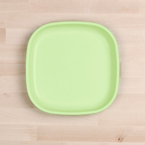 A Re-Play Flat Plate from REPLAY, crafted in a square shape with rounded corners and featuring a smooth finish, sits on a light wooden surface. This green plastic plate is made from recycled materials, embodying the principles of sustainable tableware.