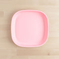 A square Re-Play Flat Plate in light pink with slightly rounded edges is centered on a light wood surface. Crafted using recycled materials, the clean, minimalist design of the REPLAY tableware emphasizes its pastel hue against the neutral background.