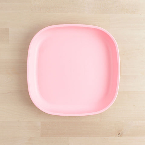 A square Re-Play Flat Plate in light pink with slightly rounded edges is centered on a light wood surface. Crafted using recycled materials, the clean, minimalist design of the REPLAY tableware emphasizes its pastel hue against the neutral background.