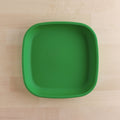 A Re-Play Flat Plate from REPLAY, characterized by its square shape and green color, is made from recycled plastic materials and positioned on a light wooden surface. The plate features rounded edges and a smooth, flat bottom, exemplifying sustainable tableware.
