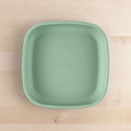 A square, light green Re-Play Flat Plate from REPLAY, crafted from recycled materials, is placed on a light wooden surface. The plate has slightly rounded edges and a smooth texture. This sustainable tableware rests on a wooden surface with a subtle grain pattern.