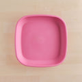 A Re-Play Flat Plate from REPLAY, featuring a pink, square design with rounded corners and made from recycled materials, is placed on a light wooden surface. The plate is empty and photographed from above.