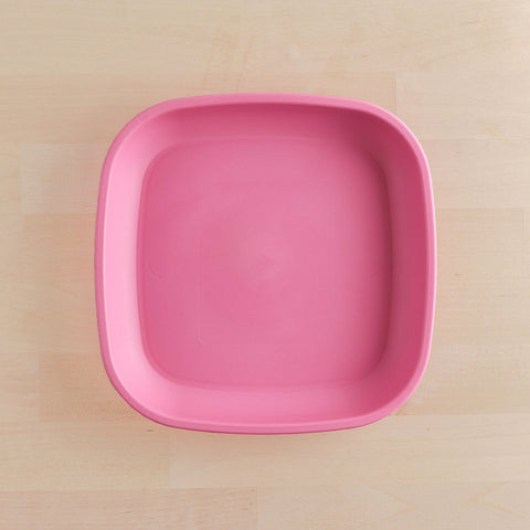 A Re-Play Flat Plate from REPLAY, featuring a pink, square design with rounded corners and made from recycled materials, is placed on a light wooden surface. The plate is empty and photographed from above.