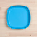 A Re-Play Flat Plate from REPLAY, featuring a bright blue color and rounded edges, is placed on a light wooden surface. This sustainable tableware, crafted from recycled materials, has a smooth glossy finish that stands out against the natural wood background.