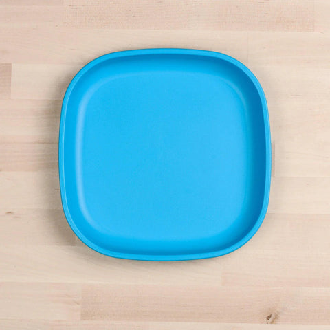 A Re-Play Flat Plate from REPLAY, featuring a bright blue color and rounded edges, is placed on a light wooden surface. This sustainable tableware, crafted from recycled materials, has a smooth glossy finish that stands out against the natural wood background.