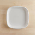 An overhead view of a simple, white, square-shaped Re-Play Flat Plate by REPLAY, made from sustainable tableware materials, is placed on a light wooden surface. The plate has slightly rounded corners.