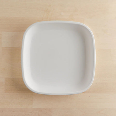 An overhead view of a simple, white, square-shaped Re-Play Flat Plate by REPLAY, made from sustainable tableware materials, is placed on a light wooden surface. The plate has slightly rounded corners.