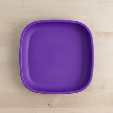 The Re-Play Flat Plate from REPLAY is a square, purple plate with rounded edges, situated on a light wood surface. The plate features a plain, matte finish without any visible designs or patterns. It exemplifies sustainable tableware, as it is crafted from recycled materials for an eco-friendly choice.
