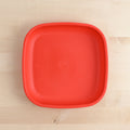 A Re-Play Flat Plate from REPLAY, characterized by its bright red color and square shape with rounded edges, is placed on a light wooden surface. This plate, crafted from recycled materials and tailored for sustainable tableware collections, is empty and features a simple, smooth design.