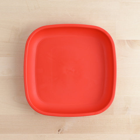 A Re-Play Flat Plate from REPLAY, characterized by its bright red color and square shape with rounded edges, is placed on a light wooden surface. This plate, crafted from recycled materials and tailored for sustainable tableware collections, is empty and features a simple, smooth design.