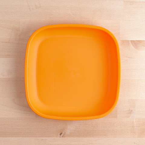 The Re-Play Flat Plate by REPLAY, an orange plastic plate made from recycled materials with slightly rounded corners and a smooth, flat base, rests on a light wood surface that displays a natural grain pattern.