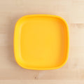 A square, bright yellow Re-Play Flat Plate by REPLAY, crafted from recycled plastic materials, sits on a light wooden surface. The durable plate features slightly rounded edges and a smooth finish, epitomizing sustainable tableware.