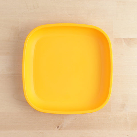 A square, bright yellow Re-Play Flat Plate by REPLAY, crafted from recycled plastic materials, sits on a light wooden surface. The durable plate features slightly rounded edges and a smooth finish, epitomizing sustainable tableware.