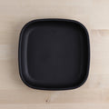 The REPLAY Re-Play Flat Plate, centered on a light wooden surface, showcases a minimalist design with rounded edges and a matte finish. Crafted from recycled materials, this black, square-shaped plate exemplifies sustainable tableware.