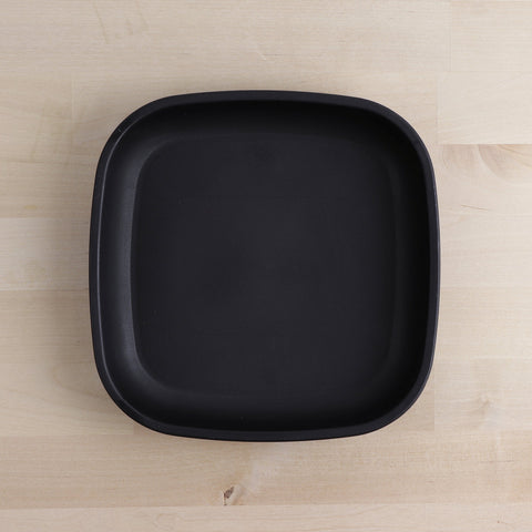 The REPLAY Re-Play Flat Plate, centered on a light wooden surface, showcases a minimalist design with rounded edges and a matte finish. Crafted from recycled materials, this black, square-shaped plate exemplifies sustainable tableware.