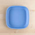 A Re-Play Flat Plate by REPLAY, featuring a square design in blue plastic made from recycled materials, sits on a light wooden surface. The plate has rounded edges and a smooth texture. This piece of sustainable tableware rests on the wooden surface, which boasts a subtle grain pattern and natural color variations.
