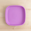 The REPLAY Re-Play Flat Plate, a square purple silicone plate made from recycled materials, sits on a light wooden surface. It features rounded edges and a smooth surface.