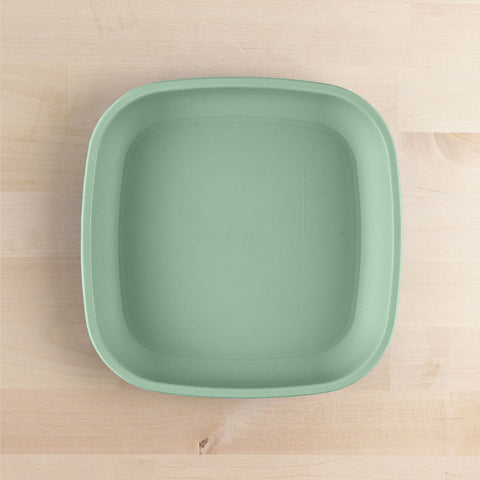 A square, light green Re-Play Flat Plate from REPLAY, crafted from recycled materials, is placed on a light wooden surface. The plate has slightly rounded edges and a smooth texture. This sustainable tableware rests on a wooden surface with a subtle grain pattern.