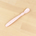 The Re-Play Infant Spoon by REPLAY, featuring a soft pink design with a long handle and a small, rounded scoop, rests on a light wooden surface. Crafted sustainably from recycled material, the handle has a slightly textured grip for improved handling.