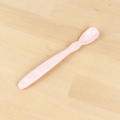 The Re-Play Infant Spoon by REPLAY, featuring a soft pink design with a long handle and a small, rounded scoop, rests on a light wooden surface. Crafted sustainably from recycled material, the handle has a slightly textured grip for improved handling.