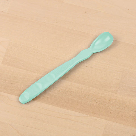 The REPLAY Re-Play Infant Spoon, in a light green color, is an elongated baby spoon crafted from recycled material. It features a small, rounded scoop on one end and a flat handle with textured grips for easy handling. Perfect for sustainable tableware collections, it rests elegantly on a wooden surface.