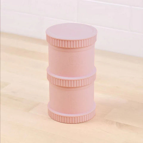 A Re-Play Snack Stack from REPLAY, featuring a pink, cylindrical design made of recycled materials, with three screw-top sections arranged vertically on a light wood surface against a tiled background. Each section includes textured grip bands and showcases a minimalist design perfect for durable snack storage.