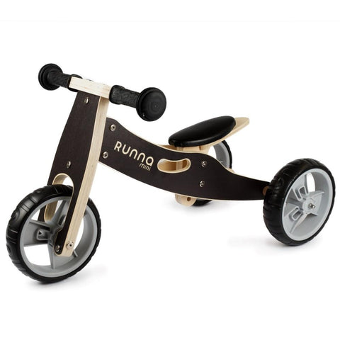 The Runna Mini - Black by RUNNA is a toddler bike designed with a wooden frame and three gray plastic wheels, perfect for beginners. This balance bike features a black seat, handlebars with black grips, and has "Runna Mini" printed on the frame, making it an ideal entry point into the world of balance bikes.