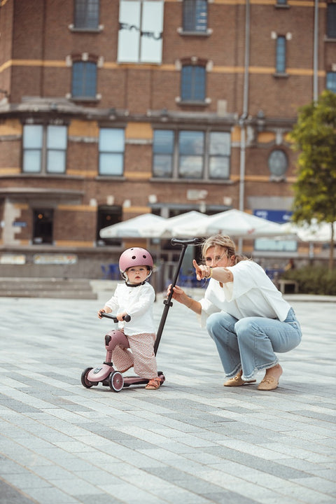A parent crouching beside a toddler learning to scoot on the Scoot & Ride Highwaykick 1 Push & Go, push bar attached