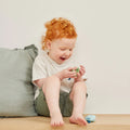 A young child with curly red hair sits barefoot on a wooden surface, near a light green cushion, examining a TIGER TRIBE Tiger Tribe Castanet made from bio plastic with a joyful expression. The child is wearing a light-colored t-shirt and green shorts against a plain, light-colored background.