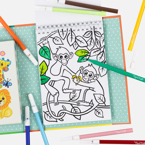 A coloring book page from the Tiger Tribe Colouring Set - Zoo by TIGER TRIBE shows two monkeys in a tree, one holding a banana. Colored markers and a children's book featuring a lion on the cover are nearby. Some parts are colored green, and the background features turquoise polka-dot paper. Perfect for kids' creative play!