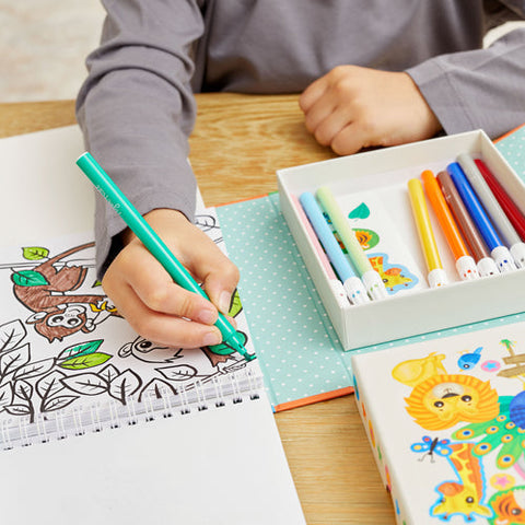 A child coloring in a book featuring a picture of an owl and leaves uses a green marker from their Tiger Tribe Colouring Set - Zoo by TIGER TRIBE, with a variety of colorful markers nearby. The scene includes the set's decorative box adorned with animal illustrations, perfect for kids' creative play.