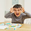 A young boy with short, dark hair is seated at a table making a playful and fierce face, his hands raised like claws. Surrounded by colorful drawing materials from his Tiger Tribe Colouring Set - Zoo by TIGER TRIBE, including crayons and a coloring book, he revels in kids creative play in a cozy room with soft decor.
