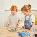 Two young children are engaged in an interactive memory challenge with the Tiger Tribe Memory + Bingo - Animals game on a wooden table. The boy with curly red hair wearing a grey T-shirt is picking up a card, while the girl with light brown hair in a ponytail, wearing a floral T-shirt and denim overalls, looks on.