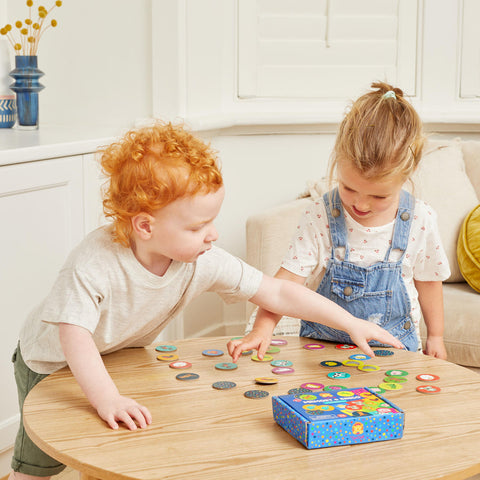 Two children are playing a game with colorful round tokens on a wooden table. The child on the left, with curly red hair, is pointing at some tokens, while the child on the right, with blond hair in a ponytail, is looking down at them. They are engaged in interactive memory challenges from "Tiger Tribe Memory + Bingo - Animals" by TIGER TRIBE. A blue game box is on the table.