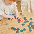 A young child with red hair is playing Tiger Tribe Memory + Bingo - Animals, an interactive game by TIGER TRIBE designed to boost cognitive skills, on a wooden table. The game consists of circular cards, some face up showing images of various animals and others face down with a polka dot pattern. The child is wearing a light gray shirt.