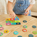 A child wearing a white shirt and denim overalls is playing Tiger Tribe Memory + Bingo - Animals on a wooden table. The game pieces, featuring colorful animal illustrations, are being placed by the child onto a square grid board. This interactive memory challenge from TIGER TRIBE enhances the child's cognitive skills.