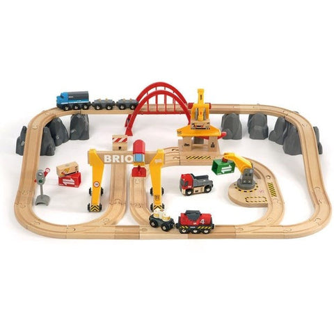 The Brio World Cargo Railway Deluxe Set by Brio includes a wooden toy train, a red bridge, multiple tracks, a crane, and magnetic vehicles such as a fire truck and various construction vehicles. The setup also features wooden mountains and tunnels, offering an immersive miniature construction-themed play experience.