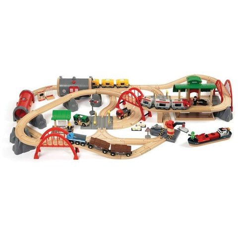Experience the Brio World Deluxe Railway Set—an expansive, intricate wooden train set featuring diverse tracks, tunnels, bridges, and accessories. A vibrant assortment of Brio trains and vehicles travels along the tracks, navigating through scenic elements such as a station, mountains, and a cargo ship.
