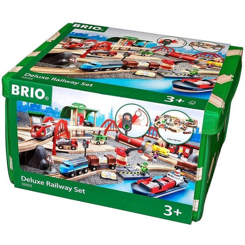 A green storage box displays an image of the Brio World Deluxe Railway Set on its sides. The set, part of the BRIO World collection, includes various train components, railway tracks, bridges, and accessories suitable for children aged 3 and up. The image depicts a colorful and detailed train scene featuring a motorized Metro city train.