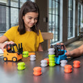 A young girl with shoulder-length dark hair plays at a table with the colorful Fat Brain Toys Forklift Frenzy set. She wears a yellow shirt and smiles while maneuvering an orange forklift from Fat Brain Toys, honing her fine motor skills. Various orange, green, and white stackable cylinders are scattered on the table.