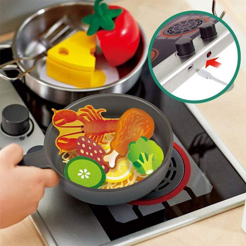 The HAPE Deluxe Kitchen with Fan Fryer from HAPE features a child's kitchen playset with an electric stove and a pan filled with toy food, including a drumstick, shrimp, vegetables, and noodles. Also on the stove are a toy cheese wedge and tomato. A close-up inset shows the power switch highlighted with a red arrow, making it perfect for imitative play.